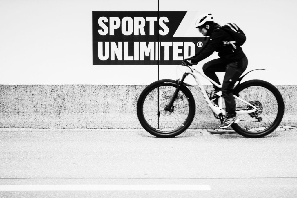 Schnappschussfotografie mit bewegtem Radfahrer vor Sports Unlimited Radfahrer fährt an einer Werbewand mit der Aufschrift Sports Unlimited vorbei
