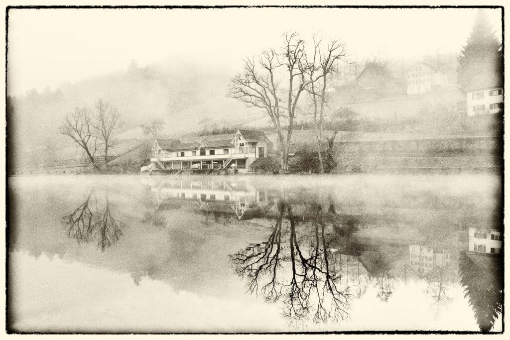 Nebel und Spiegelung erzeugen eine ruhige Schwarzweissstimmung. Nebelverhangenes Haus mit Spiegelung im See als stimmungsvolle Schwarzweissaufnahme.