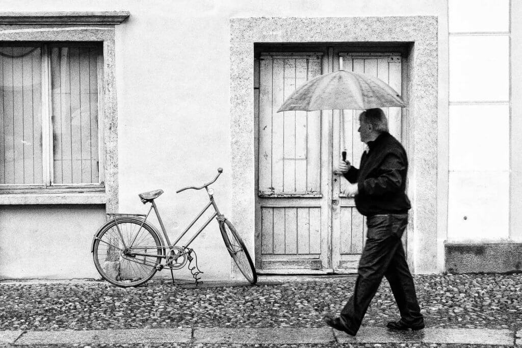 Alltagsmoment in der Streetfotografie als Schwarzweissbild. Mann mit Regenschirm geht an einem alten Fahrrad vor einer Hausfassade vorbei.